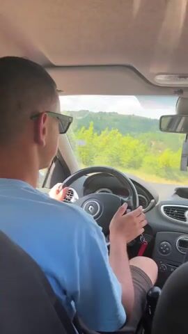 A person wearing sunglasses and a blue t-shirt is driving a car. The view is from inside the car, looking over the driver's shoulder. Outside the car, there are green hills and trees under a blue sky with white clouds. The camera also briefly shows the driver's hand on the steering wheel, the car's dashboard and rearview mirror, and the seatbelt. Another shot shows the road ahead winding through the landscape, with the passenger's arm visible out the window.