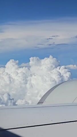 This is a short video filmed from the window of an airplane, looking out at the sky and clouds. The wing of the plane is visible in the foreground, along with part of the engine cowling. Large, puffy white clouds fill the middle ground, with a clear blue sky visible above. The sun is shining, creating bright highlights on the clouds.