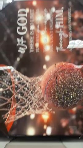 The video shows a framed piece of wall art with a basketball going through a net. The text on the art reads "With God there is no limit to what you can do. There is no obstacle you can't overcome through Him. All things are possible." The art is displayed in a retail setting, possibly a store, as evidenced by the shelving and price tags visible below the artwork. The lighting is bright, and the camera is positioned to capture the artwork from a frontal perspective, with some movement suggesting the camera operator is walking or panning.