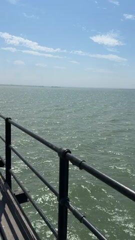The video shows a walk along a wooden pier extending out over a body of water. The pier has a black metal railing on the right side and a set of train tracks with a black metal railing on the left. The water is a greenish-blue color and appears choppy with sunlight glinting off the surface. The sky is blue with scattered white clouds. There are no people visible in the video. The camera is moving forward along the pier, providing a perspective of the path ahead and the surrounding environment. The pier is well-maintained with visible wooden planks and posts supporting the structure. A lamppost is visible on the pier in the distance. The overall impression is a bright, sunny day at the seaside.