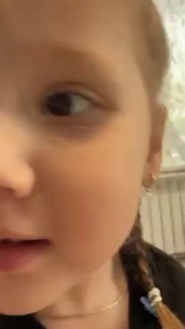 A close-up shot shows a young girl with light brown hair pulled into a braid, wearing a gold necklace and dark shirt. She has a brown mole on her left cheek and is looking at the camera with a slightly curious expression. In the background, a blurred image suggests a television or screen. Distorted music is playing in the background.