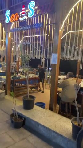 This video captures the exterior and part of the interior of an outdoor restaurant or cafe called "Sea Soul Restaurant Cafe" at night. The establishment features a prominent neon sign spelling out "Sea Soul" with a lighthouse graphic replacing the letter 'L', and "RESTAURANT CAFE" written below it.

The cafe has a rustic, open-air design with a wooden framework. Decorative elements include string lights draped across the ceiling structure and numerous ropes with small, bell-like ornaments hanging vertically, creating a unique ambiance. Potted plants are strategically placed around the dining area.

Several patrons, mostly men, are seated at various tables, which are covered with patterned tablecloths. The seating consists of wooden benches and chairs equipped with blue cushions. One man wearing a striped shirt and a cap is seen relaxing and smoking a shisha pipe at a table on the right side of the frame. Another man, possibly a staff member, is visible in the center, moving between tables. The scene suggests a relaxed and casual atmosphere, typical of an evening outing at a cafe or restaurant in Dahab, Egypt.