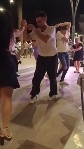 This is a video clip of a group of people dancing outdoors at night. The ground appears to be paved, and there are some lights and trees visible in the background. The people are dressed in casual attire, and some are dancing in pairs. The camera is handheld and appears to be recording from within the group. There are several individuals visible throughout the clip, including a woman in a black dress and high heels, a man in a white t-shirt and black pants, and a man in a white shirt and blue shorts. The dancing appears to be energetic and social.