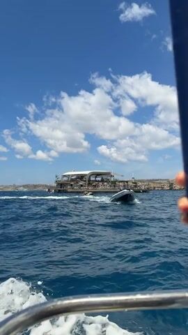 A small boat travels across deep blue water toward a large flat boat anchored near a rocky coastline. The large boat has a white roof, wooden deck, and a bar. A smaller boat circles it. The sky is bright blue with fluffy white clouds. The camera is shaky, as if it is in another moving boat. The shot is taken from a low angle.