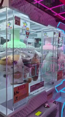 The video pans across an arcade filled with colorful claw machines and prize dispensing games. The machines are illuminated with bright pink and blue neon lights, creating a vibrant and energetic atmosphere. Many of the machines contain plush toys of various characters, some of which are packaged in plastic. The floor is a checkered pattern of black and white tiles that reflect the colorful lights. In the background, there are more rows of similar machines, suggesting a large arcade. The camera movement is smooth and steady, showcasing the visual appeal of the arcade.