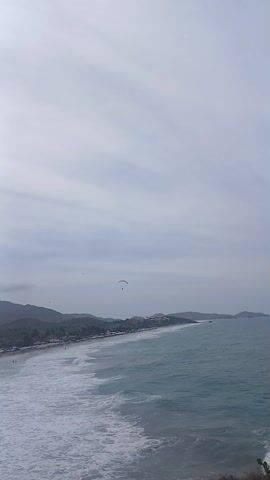 The video shows a beach on a cloudy day, with mountains in the distance and a parasailer floating above the water. The beach is populated with people, and there are several buildings along the shoreline. The ocean is a light blue color and has white waves. The camera is static, providing a wide shot of the scene.