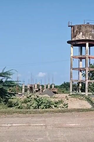 The video captures a construction site with a partially built structure and a tall, weathered water tower against a clear blue sky. Piles of sand and construction materials are visible. In the foreground, a person's hands are seen gripping the handlebars of a motorcycle, indicating movement from a rider's perspective. The motorcycle's front wheel is partially visible. The environment appears to be rural or semi-urban with vegetation surrounding the construction area.