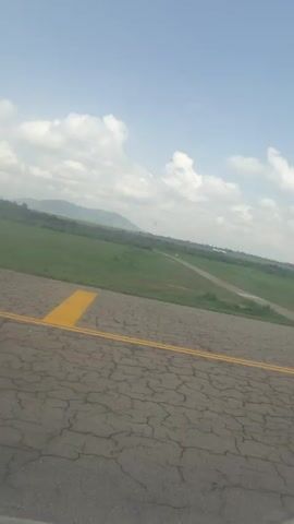 The video, shot from an elevated, forward-facing position, shows an airplane landing on a runway. The camera is positioned on a cracked asphalt surface with yellow lane markings, presumably a taxiway or apron near the runway. In the background, a grassy field slopes down towards the runway where a white and blue airplane is landing. The airplane appears to be a passenger jet. The sky is partly cloudy. Beyond the green field are some trees and distant hills. The overall environment seems to be a rural or semi-rural airport setting.