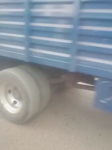 The video shows a blurred street scene from a moving vehicle. In the foreground, the side of a blue container or truck bed is visible. Beyond that, a blurry tire of a vehicle is seen. Further down the road, there are several vehicles, including a white car, a tuk-tuk, and a dark-colored vehicle. A billboard that reads "PLOTS FOR SALE" is visible behind the white car. A person in a yellow shirt is standing near the billboard. Trees and some fencing are also visible in the background. The scene appears to be a roadside market or commercial area.