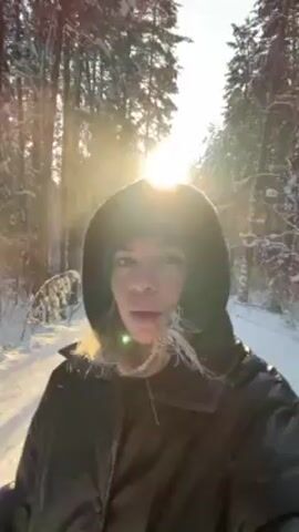 The video shows a woman standing on a snow-covered path in a forest. She is wearing a dark hooded jacket and has blonde hair. The sun is shining brightly through the trees, creating a strong backlight. In the distance, another person is skiing along the path. The environment appears to be a serene, snow-covered forest on a sunny day.