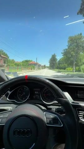 The video captures a first-person perspective from the driver's seat of an Audi car. The driver's hands are visible on the steering wheel, which is a custom carbon fiber model with red stitching and a red stripe at the top. The dashboard displays a speed of 52 km/h and other indicators. The car is driving on a paved road through a village with houses and trees on either side. The sky is clear and blue, suggesting a sunny day. The video lasts approximately four seconds.