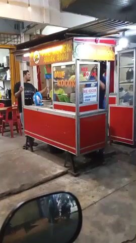 This video captures a street food stall in Bandarbireuen, Indonesia, at what appears to be nighttime or a dimly lit indoor space, possibly a market or alleyway. The primary subject is a "Mie Kocok" (a type of Indonesian noodle soup) stall, characterized by its vibrant red counter and a silver frame.

Two individuals are visible at the stall. One man, wearing a black t-shirt and jeans, stands behind the counter, seemingly interacting with the food preparation area. Another person, whose face is not clearly visible, is also behind the counter.

Prominently displayed on the stall is text in Indonesian. The main sign reads "MIE KOCONK AWAK AWAI," indicating the name of the dish and possibly the vendor. Below this, a smaller sign states "MIE KOCONK UDANG EBIE AYAM," specifying the ingredients or variations available, which include shrimp and chicken.

The scene is set against a backdrop that suggests a commercial or service area, with shelves holding various items visible in the upper left and a glimpse of more food stalls or retail spaces to the right. The ground is concrete, and the lighting appears to be artificial, coming from overhead lamps illuminating the stall.

In the foreground, a motorcycle's side mirror is visible, reflecting the scene, implying the video was recorded from a moving vehicle or from a seated position on a motorcycle. The overall impression is that of a typical evening scene at a local food vendor, offering a glimpse into everyday life and culinary offerings in Bandarbireuen.