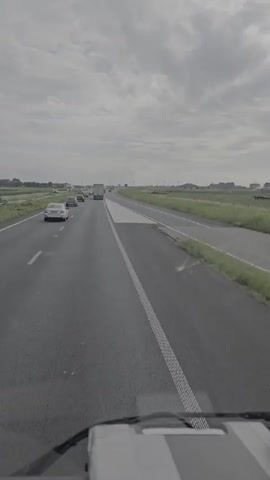 The video shows a view from the driver's seat of a vehicle on a highway. The highway has multiple lanes, and there are other vehicles on the road, including cars and trucks. The weather appears to be overcast, with a cloudy sky. The landscape on either side of the highway consists of fields and greenery. There is a distinctive white painted area on the right side of the road, possibly indicating a lane change or a merging area. The vehicle is moving forward on the highway.
