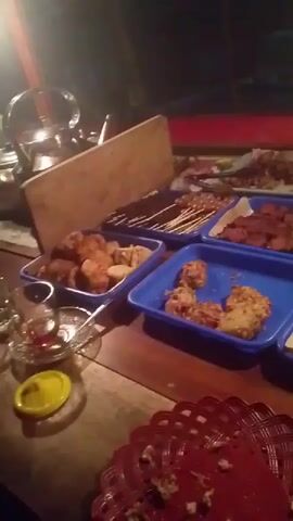 A close-up video shows various Indonesian street food dishes arranged on a wooden table. In the foreground, a red, circular, woven placemat with some crumbs is visible. To the left, a small glass of dark liquid, possibly tea, sits next to a metal utensil. Further back, there are several blue trays filled with different fried foods. One tray contains pieces of fried chicken or similar items, another appears to hold fried tempeh or tofu. Behind these, a green tray is packed with individually wrapped rice portions. The background is dark, suggesting an evening setting, with hints of red and brown surfaces. The overall impression is of a food stall or a casual dining setup with a variety of snacks and meals.