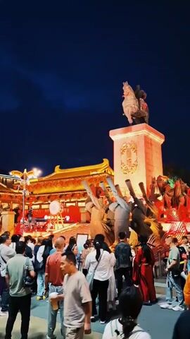 The video shows a nighttime scene in what appears to be a Chinese cultural or historical themed park. A large group of people is gathered in a public square, facing a tall pedestal on which a statue of a man on horseback stands. The pedestal is illuminated with warm light. In the background, there is a traditional Chinese-style building with a golden roof, also brightly lit.  There are additional sculptural elements around the base of the horse and rider statue, including figures with outstretched arms. People are mostly standing, looking at the scene, and some are taking pictures. The sky is dark blue. The overall atmosphere is festive and tourist-oriented.
