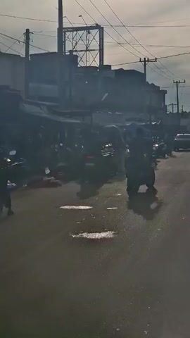The video shows a street scene with several motorcycles parked along the side of the road, some with people around them. There are also a few cars driving on the road. Buildings and power lines are visible in the background. The lighting suggests it is daytime, possibly early morning or late afternoon, with some sun glare. The road appears wet, indicating recent rain or a wet surface. The overall impression is of a busy street in a populated area.