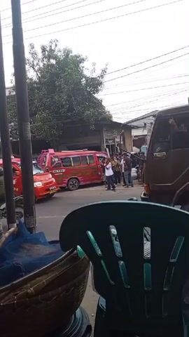 The video begins with a shot of a street scene, showing a red and pink bus and several people. The camera then pans to a market stall with various items on display, including what appear to be soccer balls wrapped in plastic, brooms, and a basket with a blue net. Several people are gathered around the stall, some appearing to be customers and others vendors. The overall atmosphere seems busy and bustling. The video is shot from a low angle, suggesting the camera is positioned on a table or a similar surface.