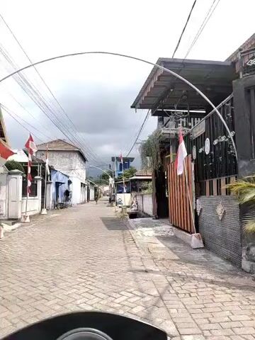 The video shows a street scene in what appears to be a village or town. The street is paved with cobblestones and lined with buildings on both sides. There are several Indonesian flags displayed, suggesting a patriotic atmosphere or a national holiday. Overhead, numerous power lines crisscross the sky, creating a somewhat cluttered visual. A person can be seen walking down the street in the distance. The sky is overcast with dark clouds, indicating potential rain or a stormy day. The architecture of the buildings is simple, with some appearing older and weathered, while others seem newer. Greenery, possibly plants or vines, is visible on some of the structures. A small drain or culvert runs along the side of the road. The perspective suggests the video is being filmed from a moving vehicle, likely a motorcycle, given the low vantage point and the slight camera shake. There are no overt signs of conflict or distress.