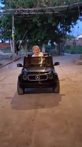 A baby sits in a black toy car. The baby is bald and wearing a white shirt and a necklace. The toy car is a Mercedes-Benz G-Wagon replica. The scene appears to be outdoors, with trees and a building in the background. The lighting suggests it is dusk or early evening.