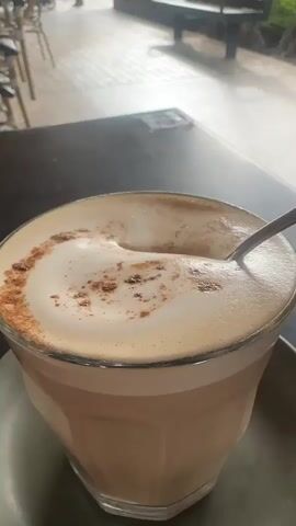 A spoon is being used to stir the foam on top of a latte in a glass. The latte has a layer of foam with some scattered cinnamon. The glass is on a dark saucer, which is on a dark table. In the background, there are blurred outdoor elements, including tables, chairs, and a person walking.