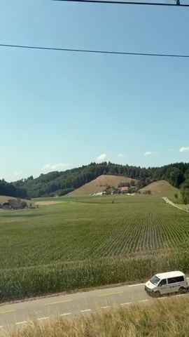 The video shows a rural landscape with rolling hills covered in trees and fields. There are several houses scattered throughout the scene, some of which appear to be farms. A road runs along the bottom of the frame, and a white van is visible on the road. The sky is clear and blue, and the sun is shining. The video appears to be filmed from a moving vehicle.