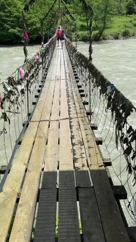 A daytime video captures a rustic wooden suspension bridge spanning a wide, fast-flowing river. The bridge features a plank walkway and metal wire mesh railings on both sides. Numerous colorful ribbons, fabric strips, and possibly other small tokens are tied along the bridge's railings, adding a decorative or traditional element.

Two people are visible walking across the bridge away from the camera, heading towards the opposite bank. The river water has a light, possibly milky green or grey hue, suggesting sediment. Lush green, tree-covered mountains or hills rise on both sides of the river, forming a scenic backdrop. The weather appears sunny, casting shadows from the bridge structure onto the walkway. The overall setting is natural and vibrant.