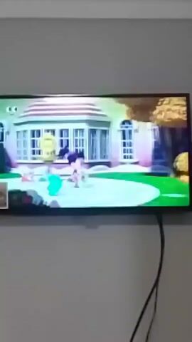 A television screen displays an animated scene. In the center of the screen, a young girl with dark hair and a turquoise top stands in a garden, holding a pink flower in her left hand and a yellow flower in her right. To her left, a young boy with dark hair wearing a brown shirt and red shorts is shown near a blue, bird-like creature. Behind them, a stylized house with white trim and large windows is visible. The ground is green, with a circular pink pathway. To the right of the girl, a large, fluffy brown object resembles a bush or a small tree. The background shows a white fence and more greenery.