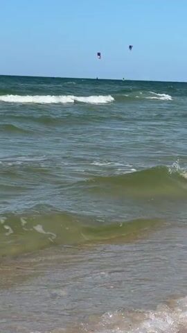 The video captures a wide shot of a beach with a clear blue sky. The ocean is a dominant feature, with waves rolling towards the shore. In the distance, two kitesurfers are visible, their colorful kites contrasting with the blue of the sky and sea. The foreground shows the sandy beach and the edge of a wave breaking. The overall scene is one of a sunny day at the beach with recreational water sports taking place.