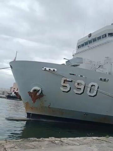 The video shows a close-up view of a gray naval ship docked at a concrete pier. The ship has the number "590" painted on its bow. The ship is large and appears to be a military vessel. The sky is overcast, and the water is calm. There is a bollard on the pier. The video pans upward to show more of the ship's structure. There is a person in the bridge of the ship, near the top.