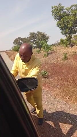 The video shows a bald man in a yellow outfit standing on a gravel road next to a black vehicle. He is attempting to reattach the side mirror of the car, which appears to be detached. The surroundings consist of dry, brown vegetation, scattered trees, and a clear sky. The camera is inside the car, providing a close-up view of the man and the mirror repair attempt.
