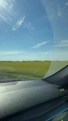 This media file is a first-person perspective video from inside a car. The scene is a rural landscape in Ulukulevo, Russia, with a vast field of bright yellow flowers, likely rapeseed, stretching across the horizon. The sky is a vibrant blue with scattered white clouds, suggesting a clear and sunny day, possibly late morning or early afternoon.

The interior of the car is visible, including the dashboard, windshield, a side window, and a rearview mirror. The windshield has some smudges and streaks. The side mirror reflects a road and the continuing yellow field. The overall mood conveyed is one of travel and appreciation for the natural scenery. There is no visible text in the media file.