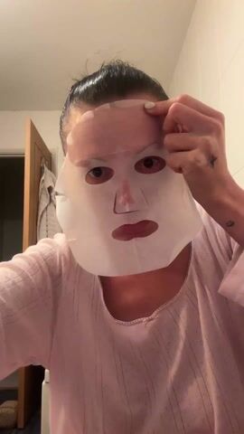 A woman with her hair pulled back and wearing a pink, striped shirt is applying a white sheet mask to her face. She is looking directly at the camera, and appears to be in a bathroom with a tiled wall and a partially visible doorway behind her. She uses both hands to adjust the mask around her eyes, nose, and mouth. She has white nail polish and small tattoos on her arms.