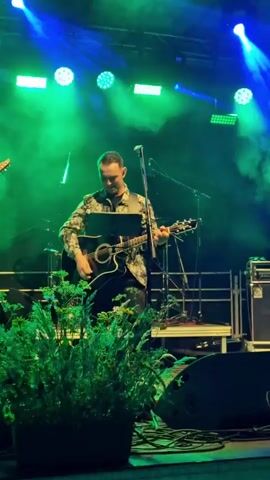 A man with short hair and a patterned shirt is playing an acoustic guitar and singing into a microphone on a stage. He is standing in front of a black music stand and is illuminated by green and blue stage lights, with fog creating a hazy effect. There are other instruments and equipment visible on the stage behind him, and a plant is in the foreground, partially obscuring the view. The overall scene suggests a live musical performance.