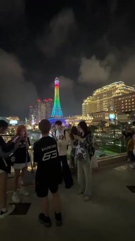 The video shows a group of people standing on a balcony or rooftop at night. They appear to be looking at and taking pictures of a brightly lit, rainbow-colored tower that resembles the Eiffel Tower. Other buildings with lights are visible in the background, suggesting an urban setting. Most of the people in the group are casually dressed and seem to be engaged in taking photos with their phones or simply looking at the view. The sky is dark with scattered clouds.