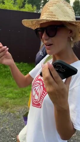 A woman wearing a straw hat, sunglasses, and a white t-shirt with a red basketball graphic is filmed outdoors. She is holding a smartphone and gesturing with her hands. Another person's arm is visible to her left, and a wooden fence and green foliage are in the background. The woman smiles and appears to be speaking.