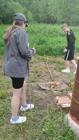 The video shows a young woman and a young man in a grassy outdoor area. The woman, seen from the back, wears a gray hooded sweatshirt, black shorts, white Crocs with blue socks, and a gray baseball cap. She holds a stick in her right hand and appears to be either lighting or has just lit something with a lighter held in her left hand. In front of her is a small fire in a makeshift fire pit made of bricks and a metal plate. The young man, wearing a black long-sleeved shirt and black shorts with white stripes, stands a few feet away from the fire, looking at it. The background features a grassy field and a line of trees. A brown barrel-like structure stands to the right of the fire pit.
