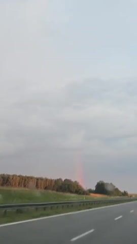 The video shows a view from a moving vehicle on a highway. On the left side of the road, there is a field of trees and some taller vegetation. A faint rainbow is visible in the distance, appearing as a vertical streak of pink light against a cloudy sky. The highway itself is paved and has white lane markings. The sky is overcast with grey clouds. No people are visible.
