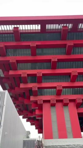 The video pans upwards, focusing on the exterior of a tall, red, multi-story building. The building features a repetitive architectural design with horizontal red beams supported by vertical red columns. Each intersection of beams and columns appears to have a decorative, square element with intricate carvings. Between the red structural elements, dark tinted glass windows or panels are visible. To the left and slightly below the main red structure, a portion of a gray, modern-looking building with vertical paneling is visible. The sky above is overcast and white. No people are clearly discernible within the frame of the video.