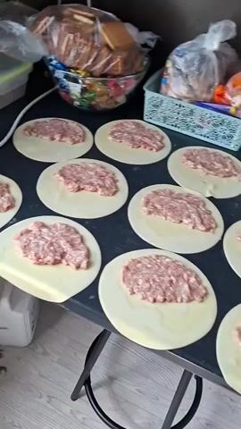 The video pans across a dark countertop where several circles of dough are laid out. Each circle of dough has a mound of raw ground meat placed in the center. In the background, there are various items including plastic bags, a clear container, bottles of green liquid, a carton of red juice, and a white salt shaker. A portion of a white appliance and black legs of a chair are visible on the left side of the frame. The countertop appears to be a dark, possibly black, surface. The lighting suggests an indoor environment. The overall scene depicts food preparation.