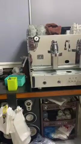 The video shows a silver, professional espresso machine sitting on a brown table. Behind the machine, there are a number of glasses and what appears to be a coffee grinder. The table underneath the machine has shelves with various items on them. The camera moves around the machine, showing it from different angles. A person in a red plaid shirt is reflected in the machine's surface. Music plays in the background.