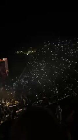 A night-time video shows a singer/pianist performing for a large audience in an arena. He is seated at a keyboard in the center of a circular, raised stage. The audience is packed into tiered seating in the darkness, many holding up illuminated mobile phones. A large display screen shows a close-up image of the performer’s face. Security personnel in neon vests are visible.