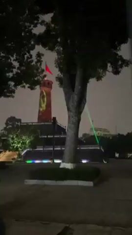 The video shows a tall, stone tower at night, illuminated with a projection of a communist hammer and sickle symbol in green and a red flag at the top. The tower is part of a larger structure with steps leading up to it. A tree stands in the foreground, partially obscuring the view. Multicolored lights line the base of the tower, and white spotlights shine from behind it. The sky is overcast, and the overall atmosphere is somewhat hazy. The camera slowly zooms in on the tower.