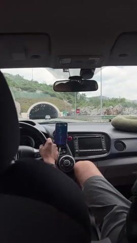 A car is entering a tunnel. The view is from inside the car looking out through the windshield. A person is driving. The driver’s hand is visible on the steering wheel and part of their bare leg is visible. The car has a phone mounted on the dashboard, presumably for navigation. There is a green pillow on the passenger seat. Inside the tunnel, there are evenly spaced lights on the roof and arrows on the walls. Another car is in front of the car in the tunnel.