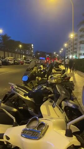 The video opens with a close-up of parked motorcycles, their chrome and paint reflecting the ambient light. The camera then pans up and to the left, revealing a street scene at dusk. Palm trees line the sidewalk, and streetlights cast a warm glow. On the opposite side of the street, cars are parked in a lot alongside a modern building. The sky is a deep blue, transitioning from twilight. Two individuals, silhouetted against the fading light, walk away from the camera on the sidewalk. The sound of distant traffic can be heard, along with a faint, rhythmic electronic beat. The overall atmosphere is calm and urban.