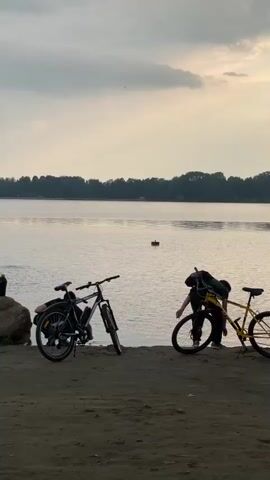 Two people and two bicycles are situated on a sandy embankment beside a large body of water. The sky is overcast with clouds. On the left, a black bicycle is parked next to a large rock. A person in a black shirt and backpack stands near this bicycle, holding a phone. On the right, a yellow bicycle is being adjusted by a person wearing a dark shirt and backpack. This person is leaning over the bicycle, appearing to work on its front wheel. In the water, two red buoys are visible. The person on the left eventually turns and walks away from the water, while the person on the right continues to tend to their bicycle.