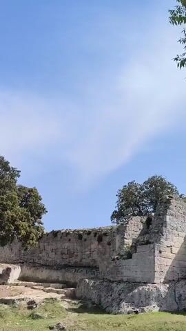 The video opens with a static shot of ancient stone ruins, featuring a prominent green tree in the foreground under a bright blue sky with wispy clouds. The camera then pans upwards, transitioning to a view of lush green treetops against the clear sky. A group of people, including several children and adults, is then shown walking along a path within the archaeological site. They are casually dressed for warm weather, with some wearing hats, and appear to be on an outing, observing their surroundings.

The camera tracks the group as they move alongside weathered stone walls and structures, highlighting the historical setting. The scene features close-ups of the visitors interspersed with wider shots that capture the scope of the ruins and the surrounding natural environment, including various trees and grasses. The video also presents a panoramic view of the expansive landscape, revealing distant hills and a valley, suggesting an elevated location. The activity seems to be a leisurely visit or tour of the historical site. The weather remains consistently bright and sunny throughout, with a clear blue sky. The setting is defined by ancient stone architecture and vibrant green foliage, indicative of a historical landmark in Olèrdola, Spain. No visible text appears in the video.