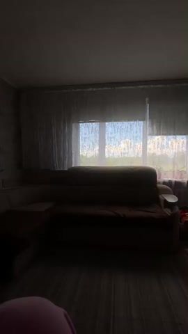 A living room is shown with a couch, windows with sheer curtains, and a TV stand. The room is dimly lit. There are toys on the floor near the windows. The camera pans across the room, revealing a wardrobe on the right side.