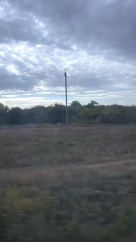 The video shows a landscape with a cloudy sky and a rural scene with houses and trees. The camera appears to be moving, possibly from a vehicle, as the foreground shows a blurred grassy area. Several buildings and fences are visible in the mid-ground, along with some vehicles. The overall impression is of a somewhat overcast day in a village or small town.