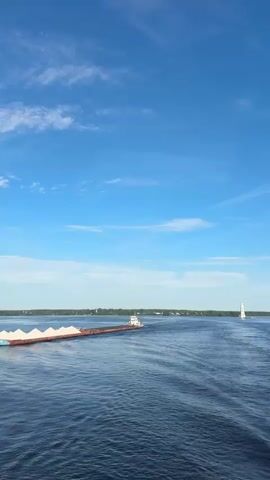 The video shows a wide, calm body of water, likely a river or lake, under a bright blue sky with scattered white clouds. In the distance, a long barge carrying what appears to be sand or gravel is moving from left to right. A small tugboat is attached to the front of the barge, pulling it. Further to the right, a white sailboat with its sail up is also visible on the water. The water has gentle ripples, suggesting a light breeze. The shoreline is visible in the far distance, appearing as a thin line of green trees and land. The overall scene is serene and depicts a sunny day with moderate water traffic.