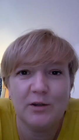 The media file is a close-up, slightly blurry selfie of a woman with short, blonde hair and a yellow shirt. She is looking directly at the camera with a neutral expression. Her face fills most of the frame, and the background is out of focus, appearing as a plain, light-colored wall. The lighting suggests it could be indoors, possibly during the daytime, given the soft light. The woman appears to be in her mid-30s to early 40s. There is no visible text in the frame. The context indicates the location is Khadyzhensk, Russia, but there are no visual cues to confirm this.